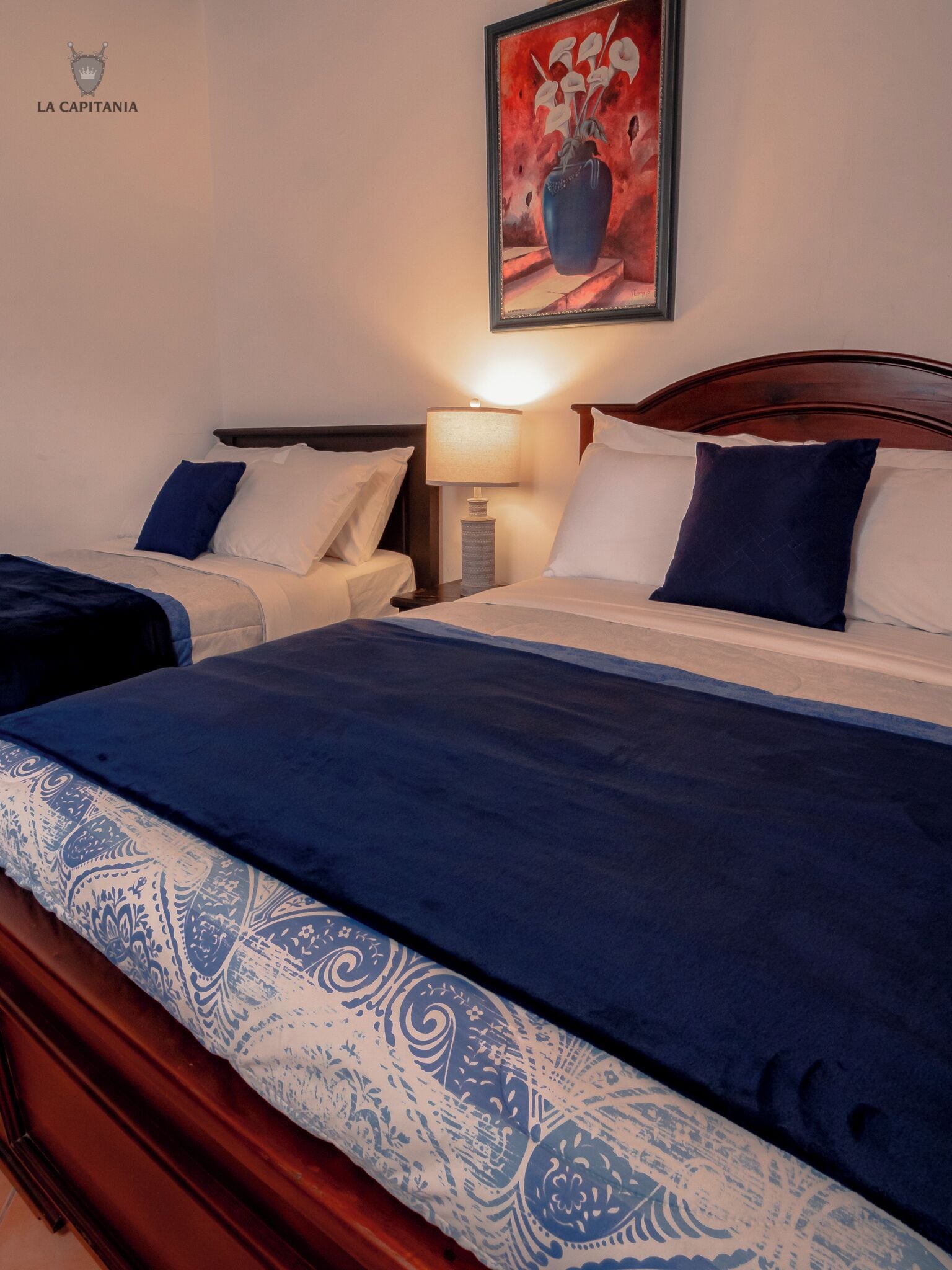 Una habitación de hotel con dos camas, una mesita de noche con lámpara y un cuadro en la pared.