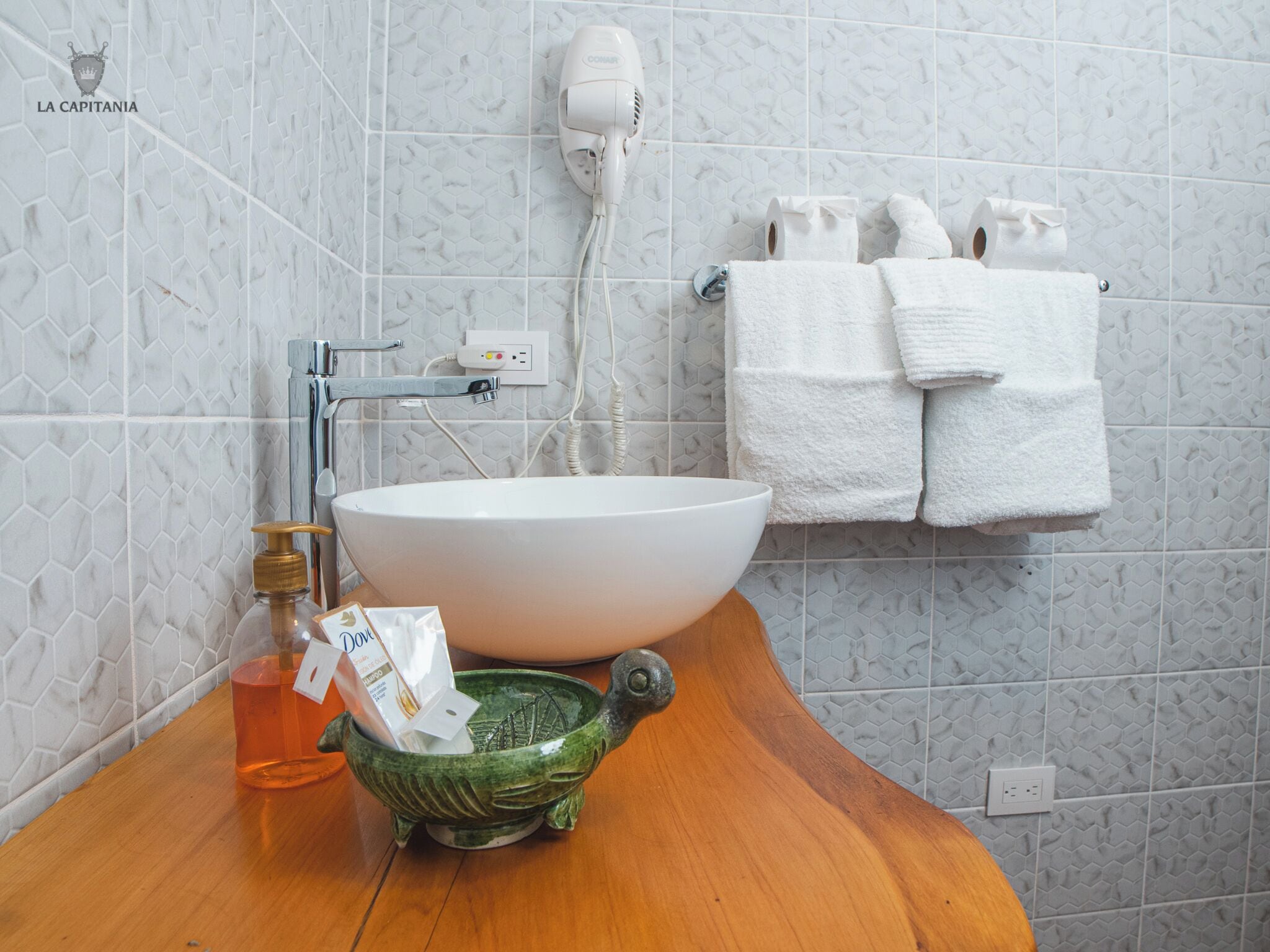 A bathroom with a white vessel sink, a hairdryer, a green ceramic bowl, and a wooden countertop.