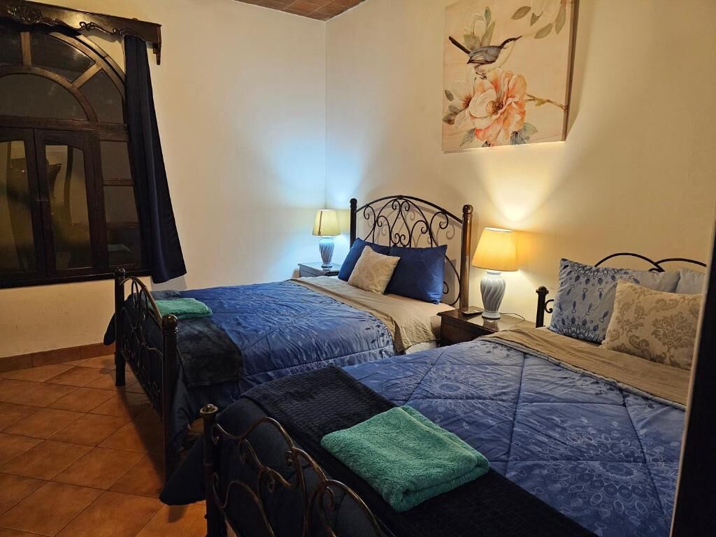 Et soveværelse med to senge, hver med et blåt quilt og et grønt håndklæde, samt et natbord med en lampe.