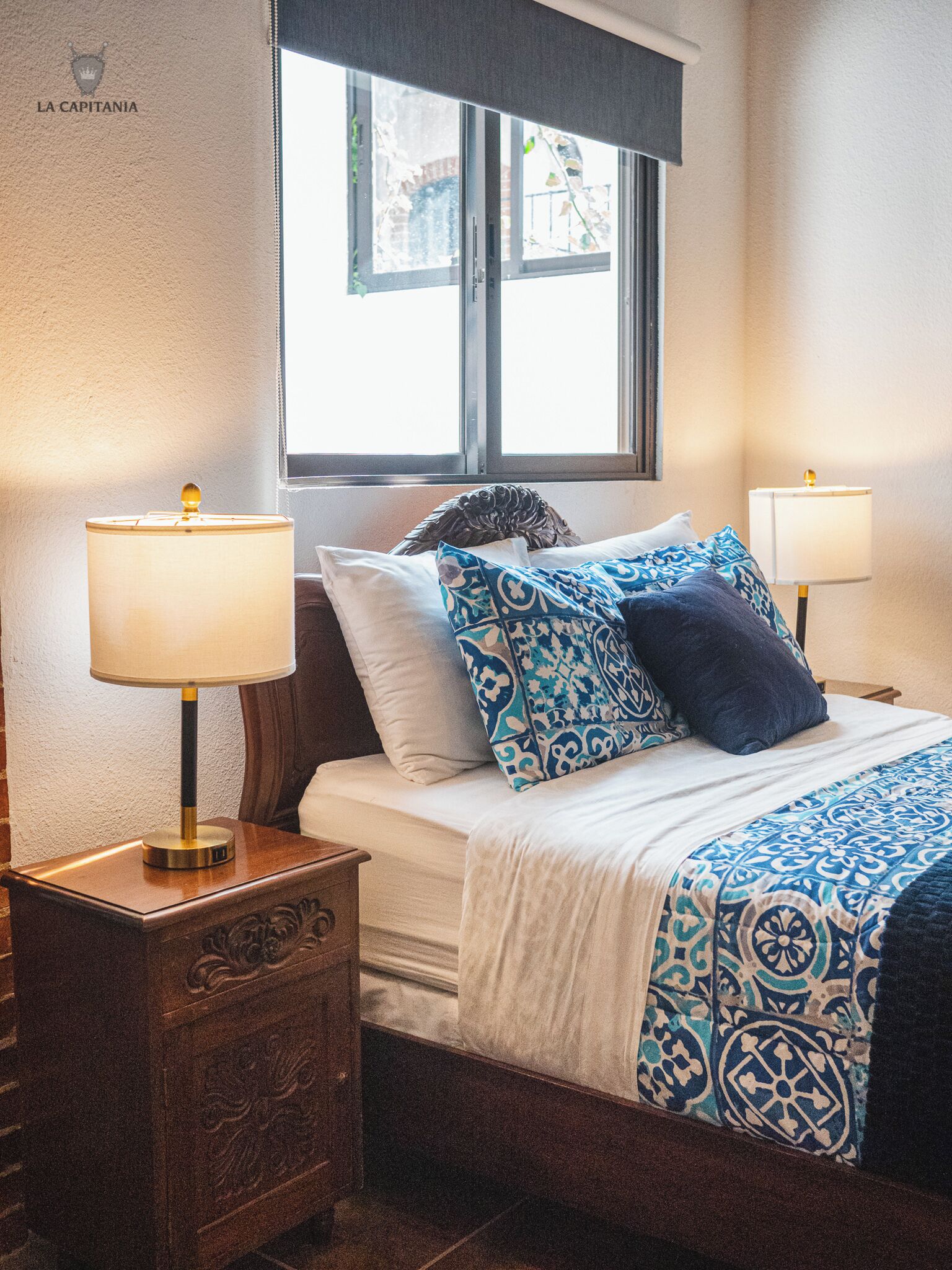 Ein ordentlich bezogenes Bett mit gemusterten Betttextilien, zwei Nachttische mit Lampen und ein Fenster mit Blick ins Freie.