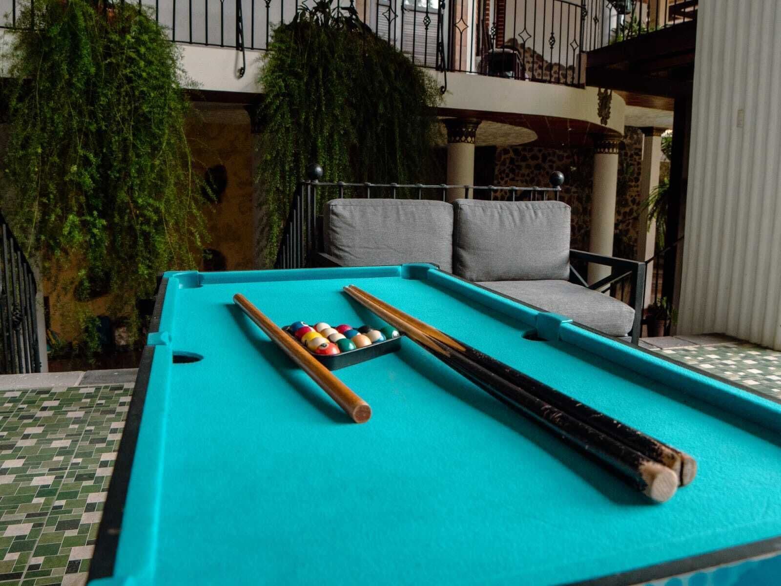 Une table de billard avec des boules et des queues, entourée de canapés gris et donnant sur un balcon agrémenté de plantes.