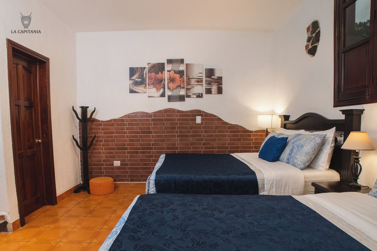A hotel room with a brick headboard, two beds, a nightstand with a lamp, a wall-mounted artwork, and a coat rack.