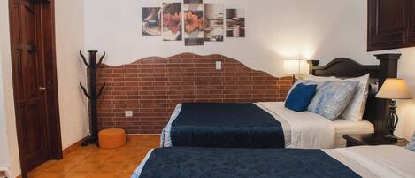 A hotel room with a brick headboard, two beds, a nightstand with a lamp, a wall-mounted artwork, and a coat rack.