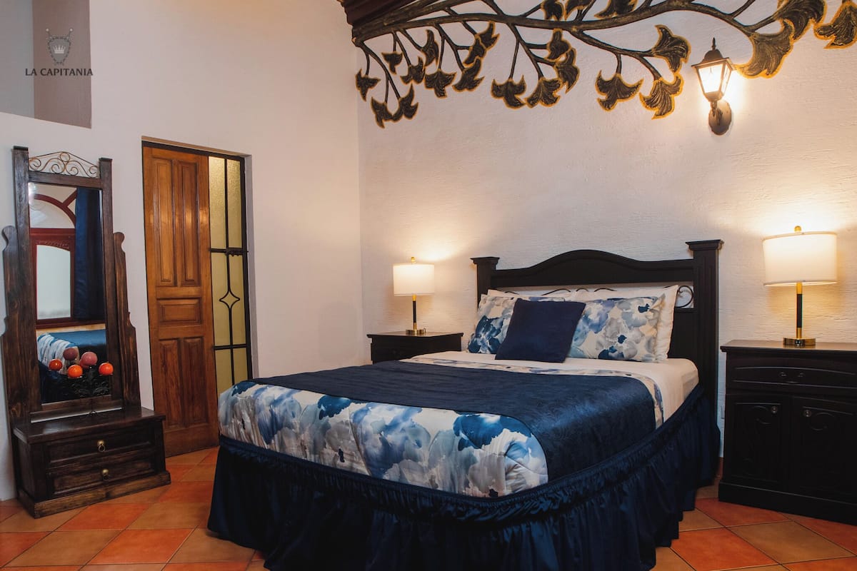 A bedroom with a bed, two bedside tables, a nightstand, a mirror, and a decorative wall piece.