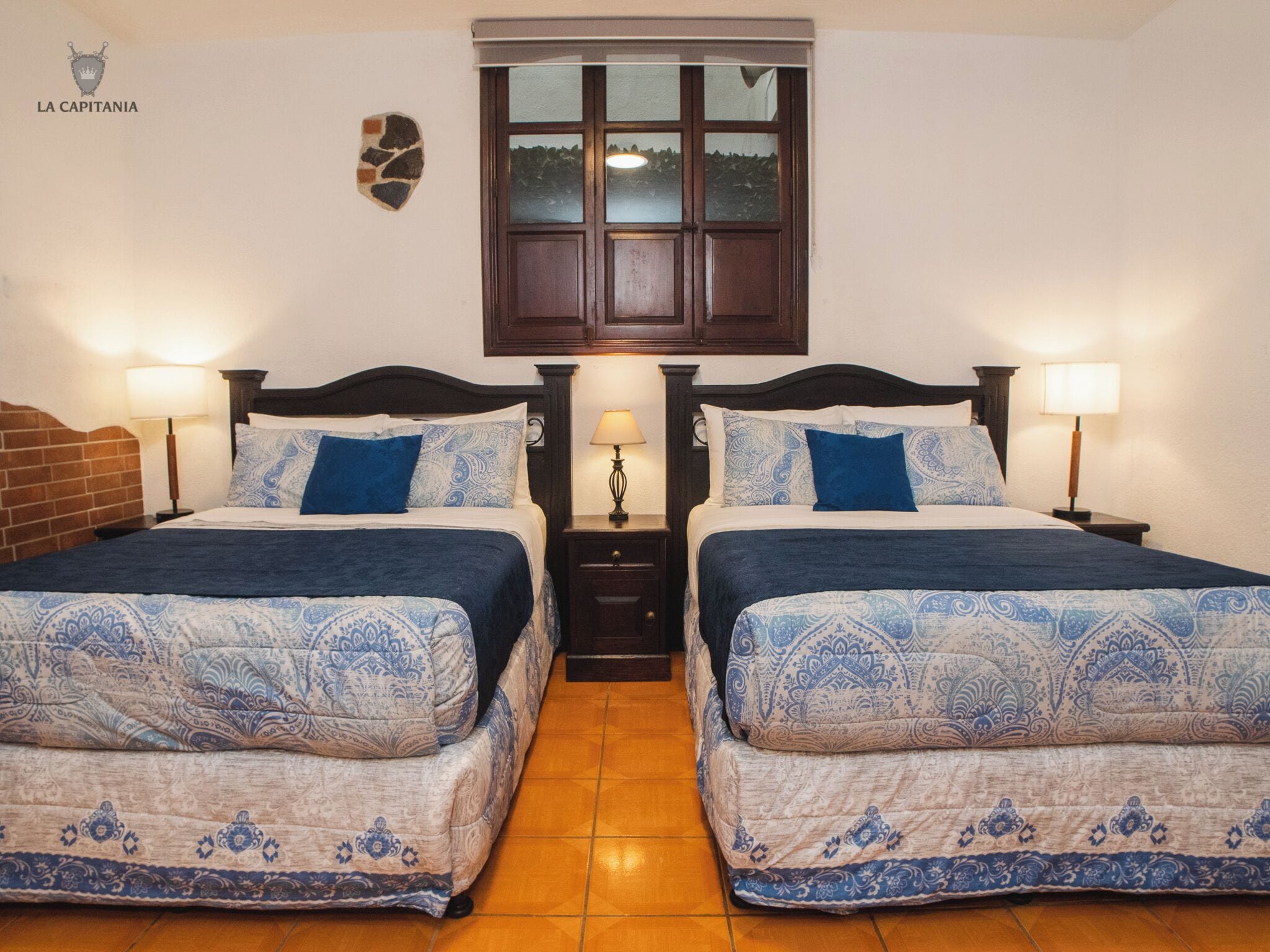 A double room with two beds, each with a headboard, bedside tables with lamps, a stone wall decoration, and a window above the beds.