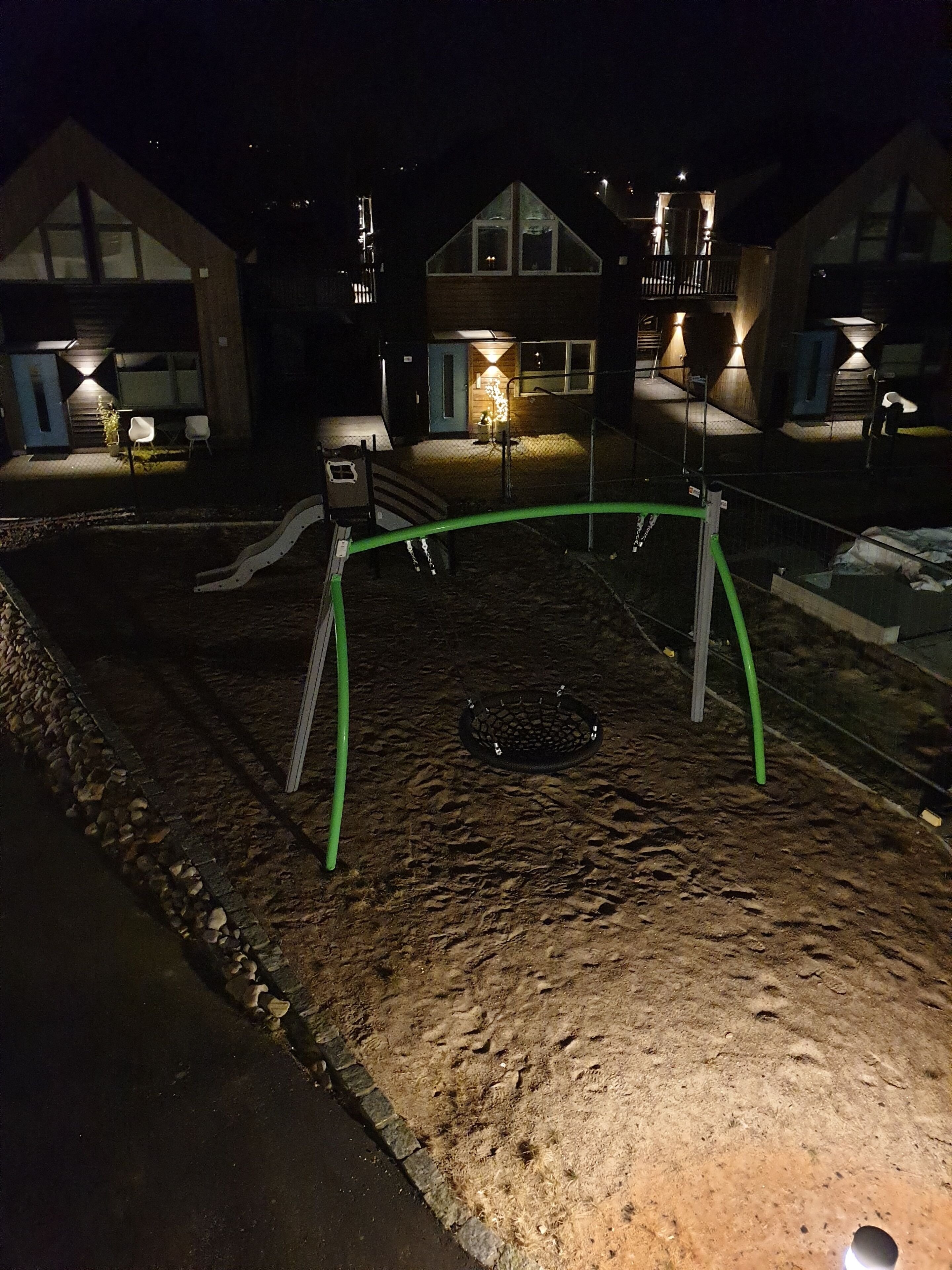 A playground with a swing set and a slide, surrounded by a paved area and a building with illuminated windows.
