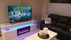 Una sala de estar moderna con un televisor de pantalla plana sobre un mueble blanco, un sofá de cuero negro con un cojín de diseño meditativo y una mesa de centro minimalista con objetos decorativos.