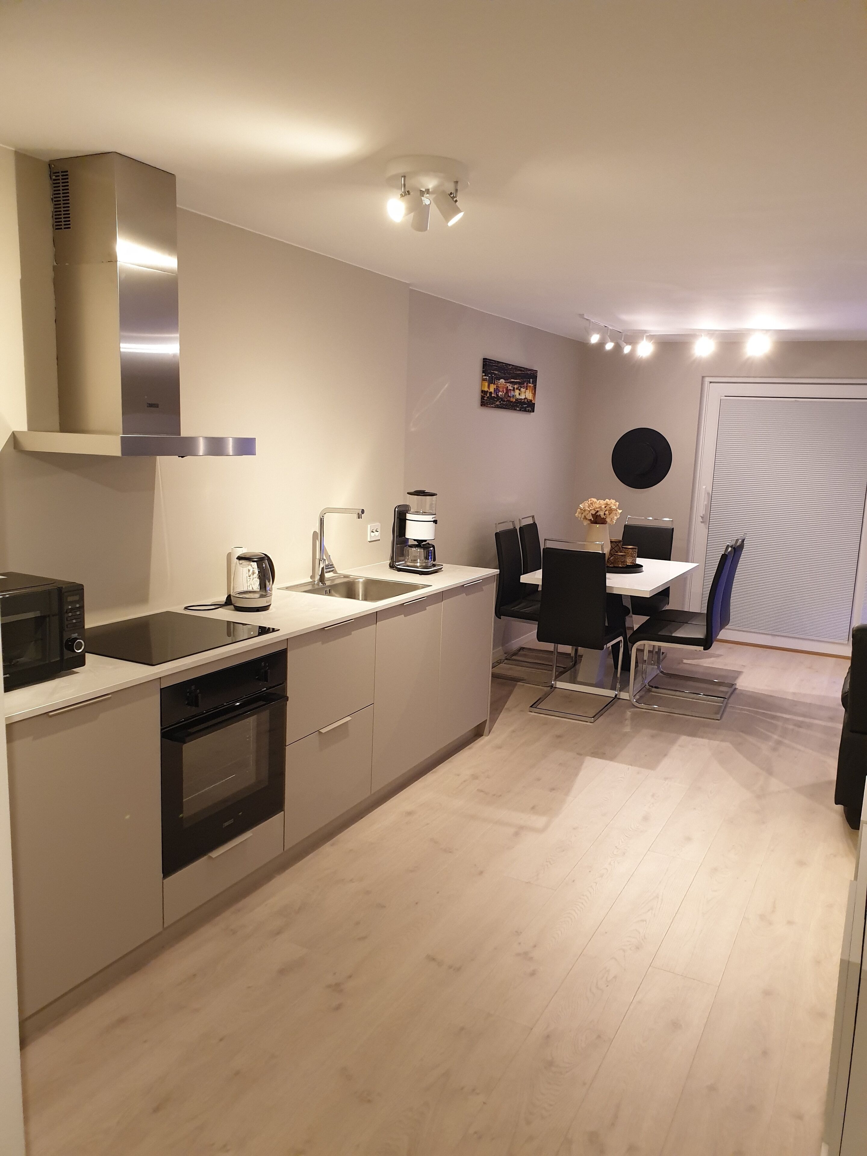 Una cocina moderna con horno empotrado, microondas y cafetera. Hay una zona de comedor con mesa y sillas, y un ventanal con persianas.