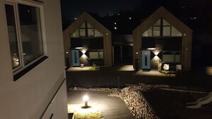 Vista nocturna de un edificio con dos puertas de garaje iluminadas, un camino bien iluminado y un pequeño jardín con un farol.