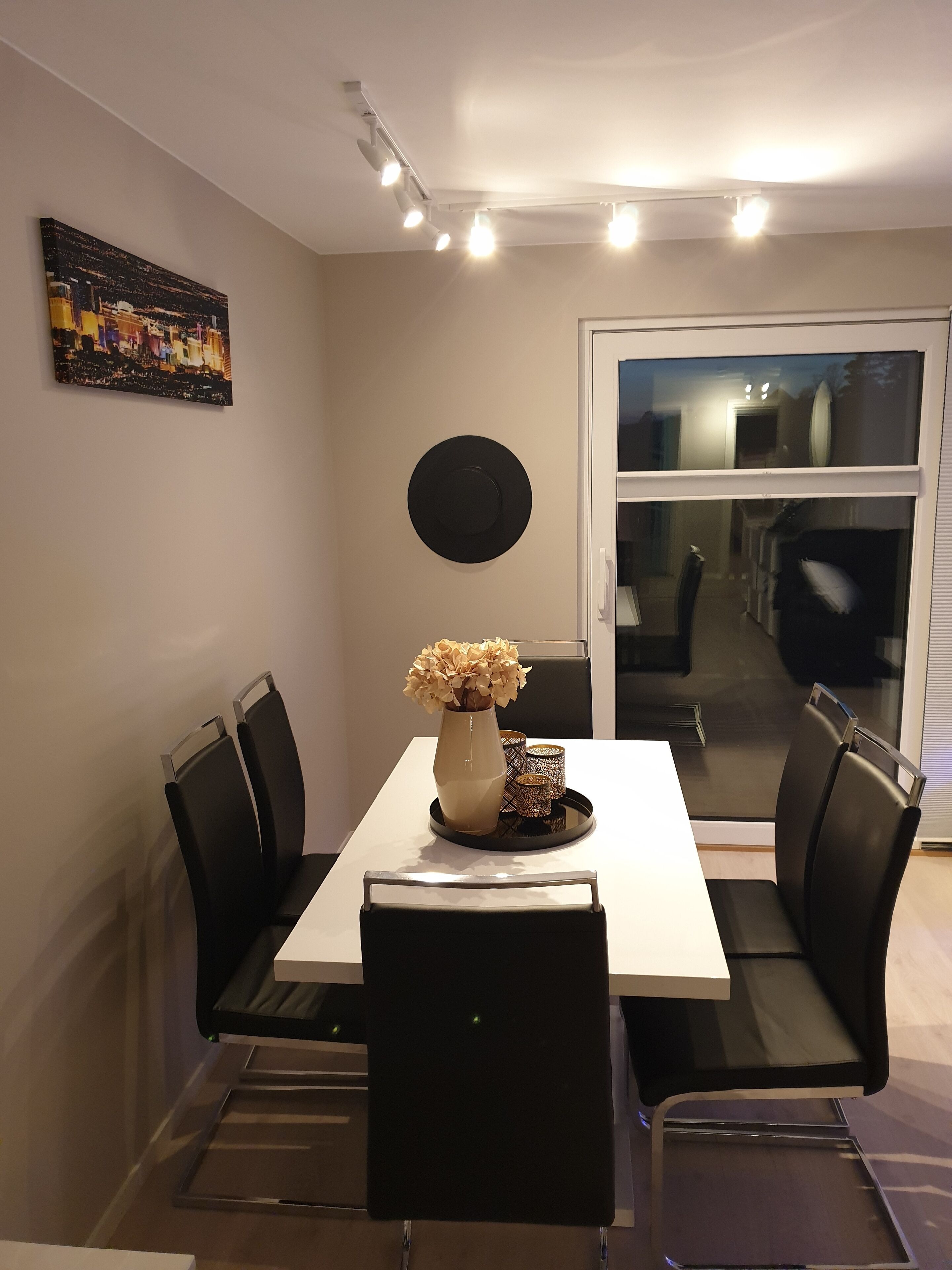 A dining area with a white table, black chairs, a vase with flowers, and a cityscape painting on the wall.