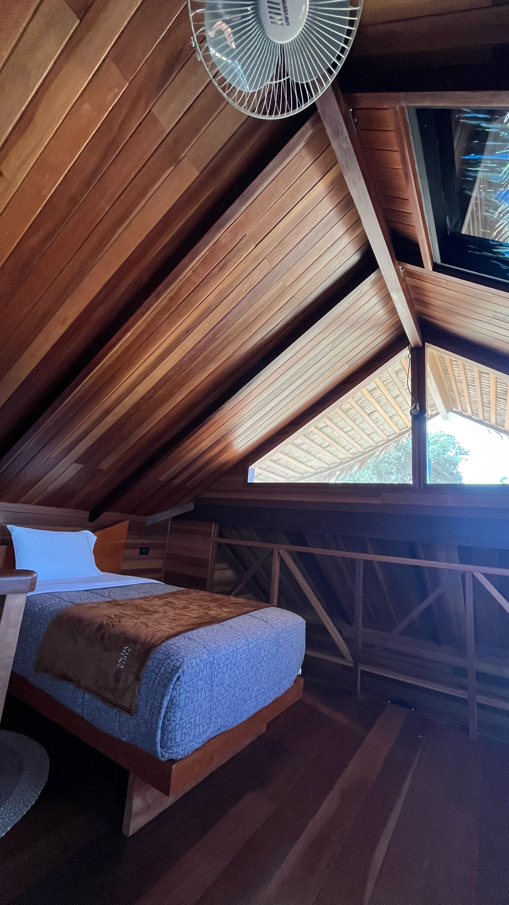 A cozy attic bedroom with a sloped ceiling, a bed with a blue patterned bedspread, a wooden headboard, and a ceiling fan.