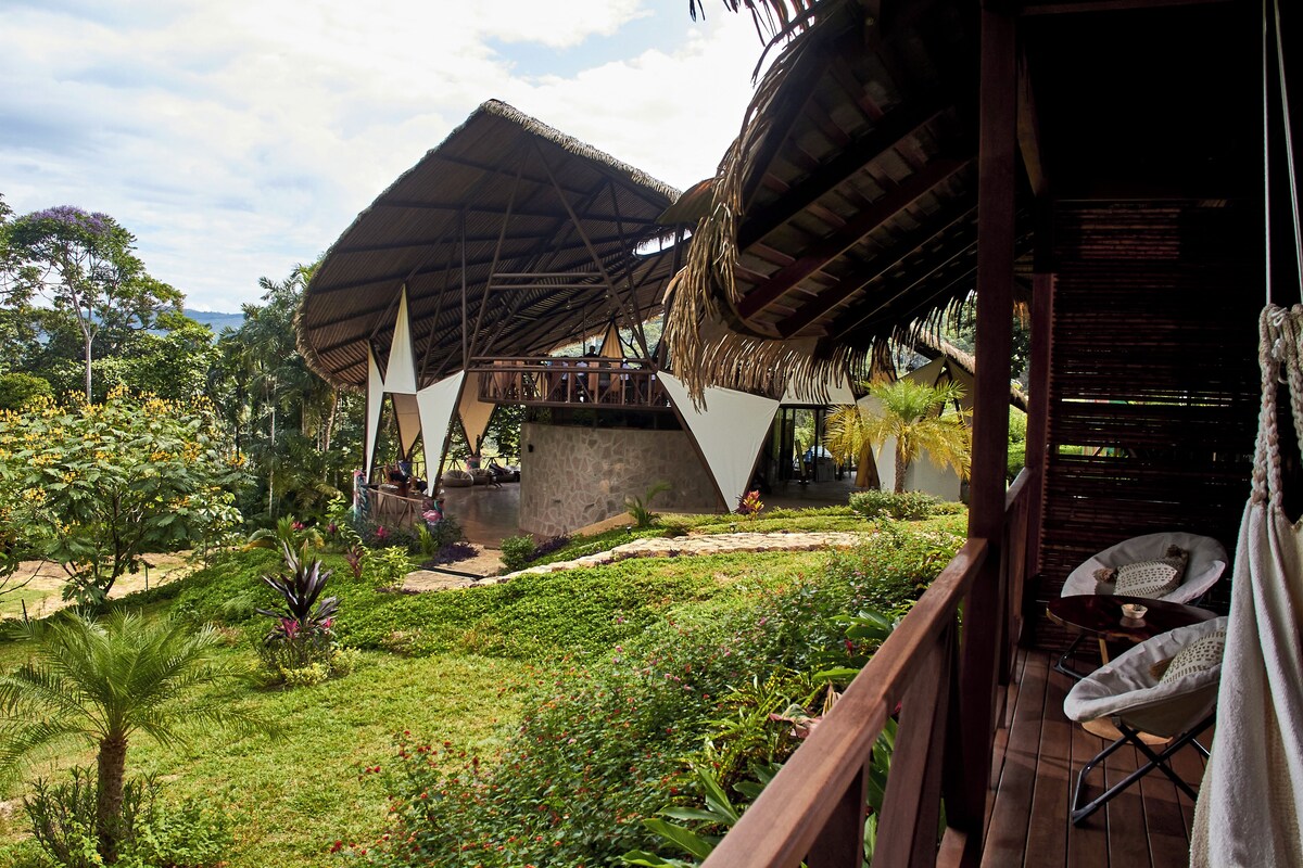 A wooden deck with a thatched-roof structure, surrounded by lush greenery and a clear sky.