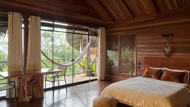 A spacious bedroom with a large bed, wooden flooring, a hammock, and a view of greenery outside. - Sachakuna Resort (Sauce)