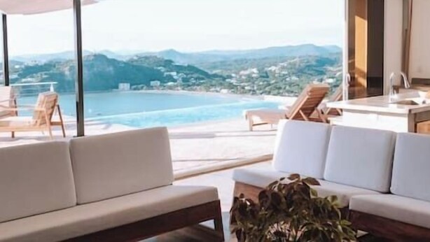 Una stanza moderna con un'ampia finestra vista piscina e montagne. Sono presenti due divani bianchi e un tavolino con sedie.