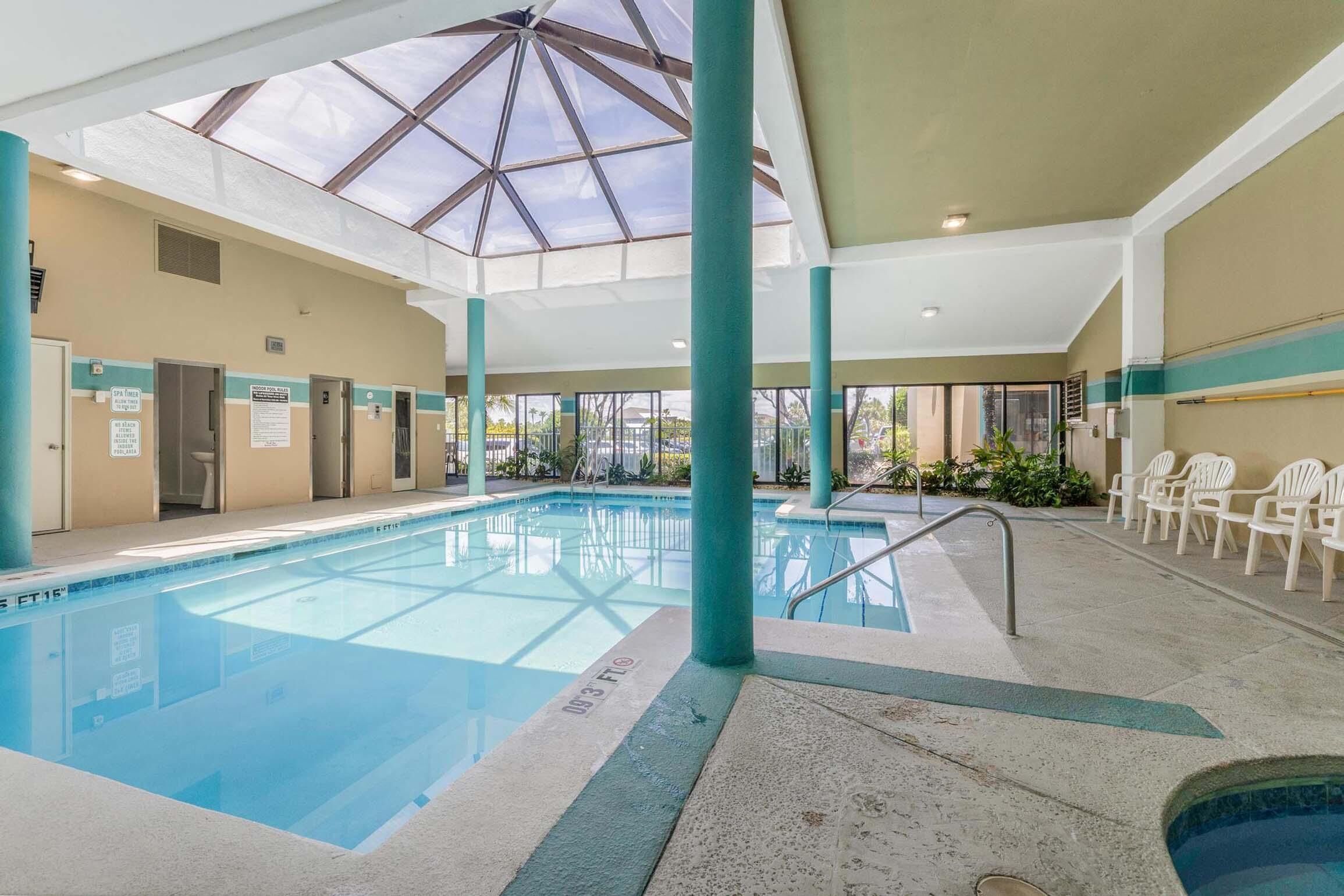 An indoor swimming pool with clear blue water, surrounded by white chairs and a glass ceiling.