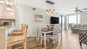 A spacious living room with a grey sofa, a dining area with a wooden table and chairs, a flat-screen TV mounted on the wall, and a ceiling fan. - Romar Place 302 (Orange Beach)