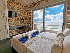 A bedroom with a stone wall, a bed with a white comforter, a blue towel, and a view of the sea through a glass door. - Pyrgos Plakias Villas (Agios Vasileios)