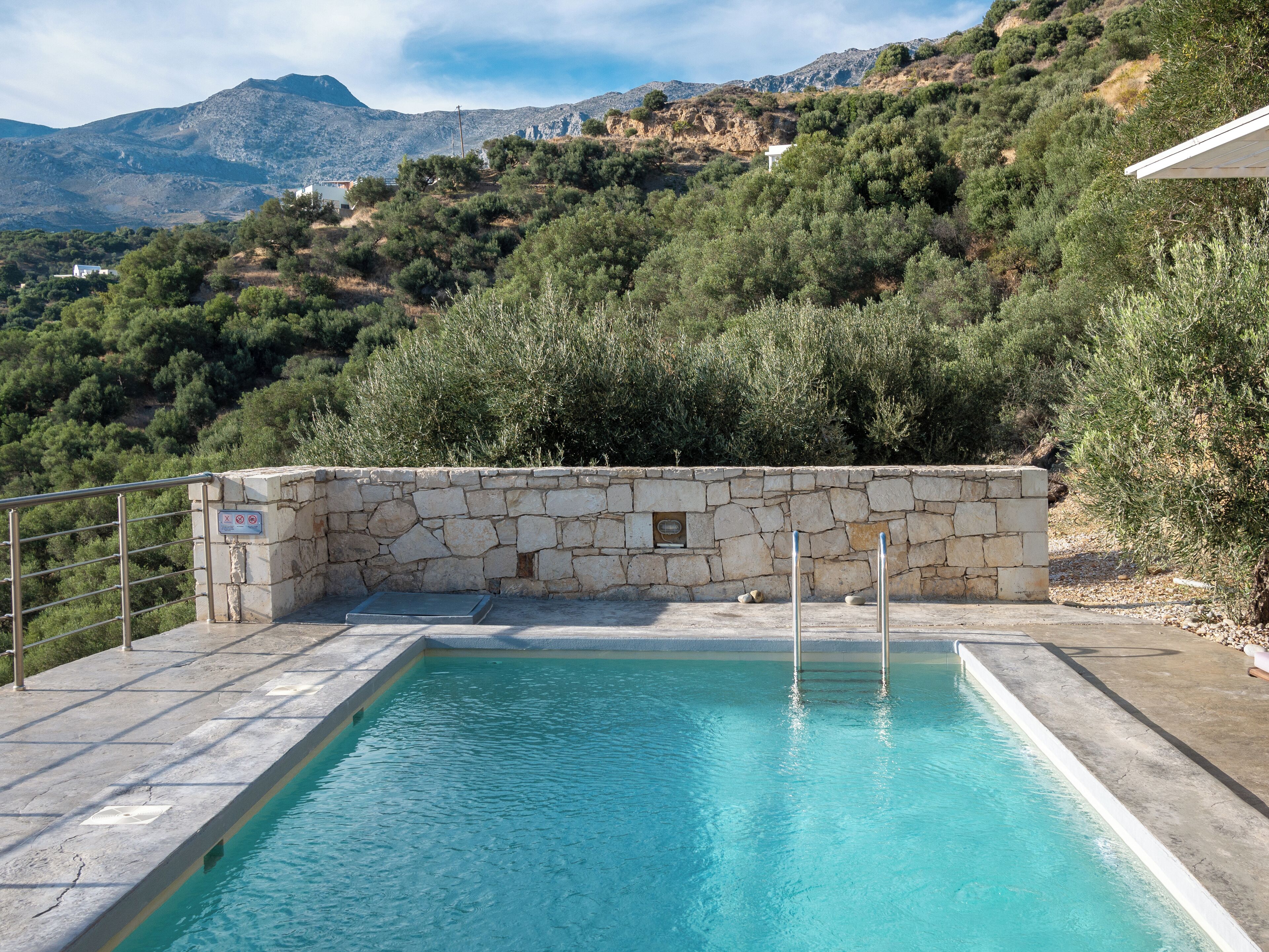 A swimming pool with stone walls and metal railings, surrounded by lush greenery and mountains in the background.