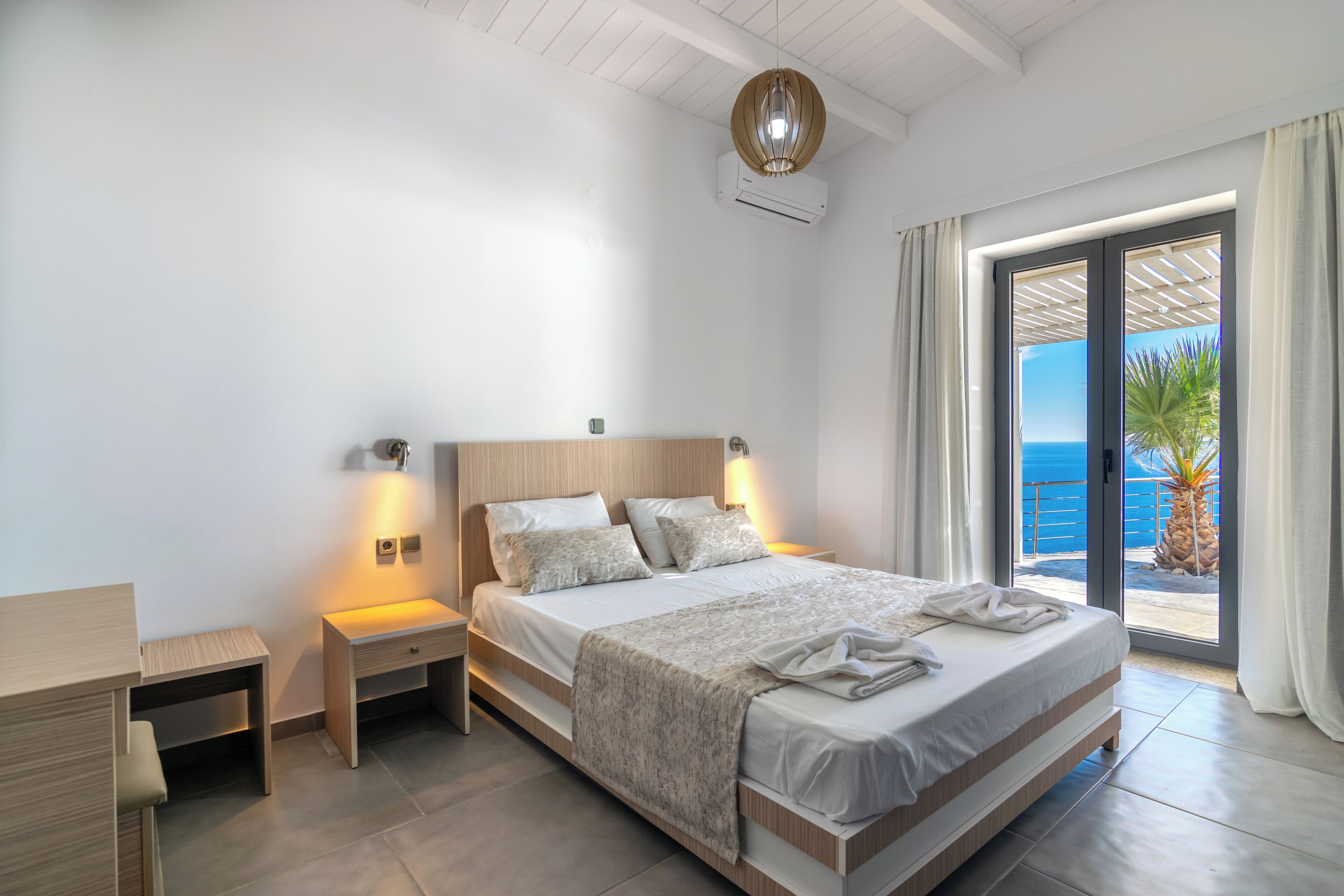 A modern bedroom with a large bed, bedside tables, a desk, and a view of the sea through a sliding glass door.
