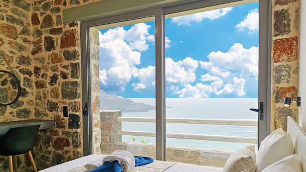 A room with a stone wall, a bed with white and blue towels, and a view of the sea and sky through a glass door. - Pyrgos Plakias Villas (Agios Vasileios)
