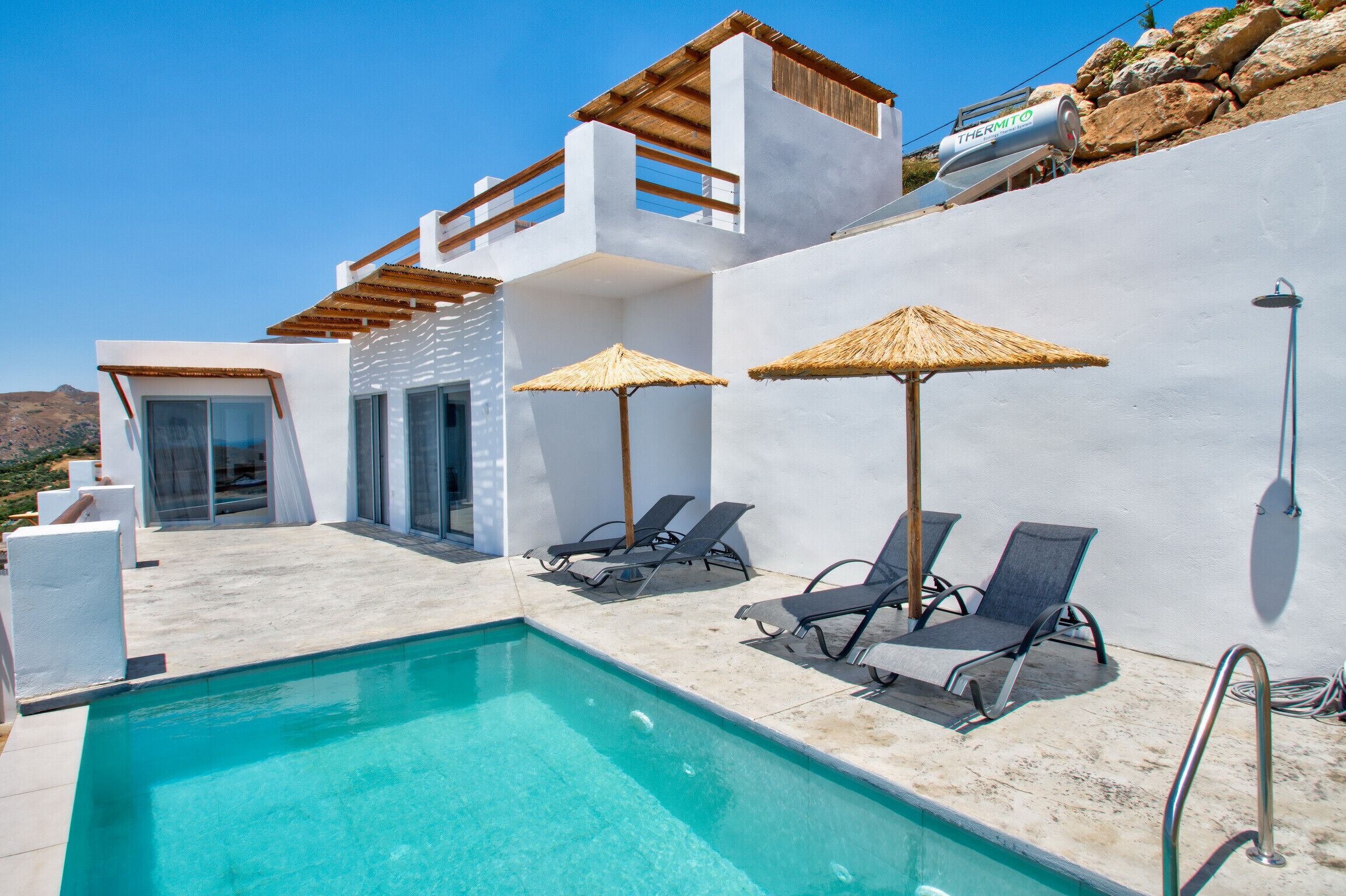 A modern pool area with lounge chairs and umbrellas, a clear blue pool, and a white building with a wooden pergola.
