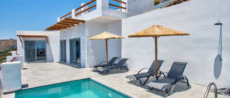 A modern pool area with lounge chairs and umbrellas, a clear blue pool, and a white building with a wooden pergola.