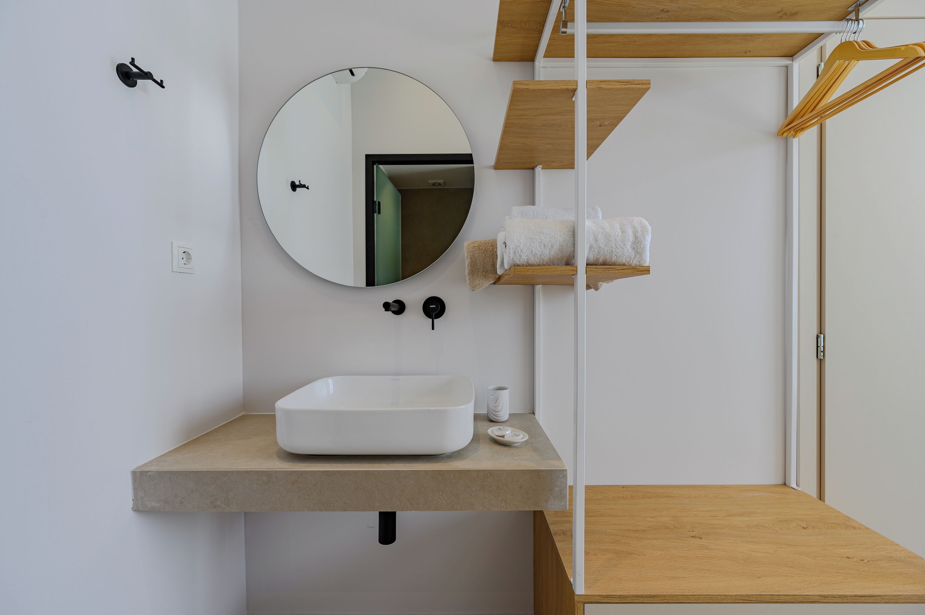 A modern bathroom with a white sink, round mirror, towel rack, and wooden shelves.