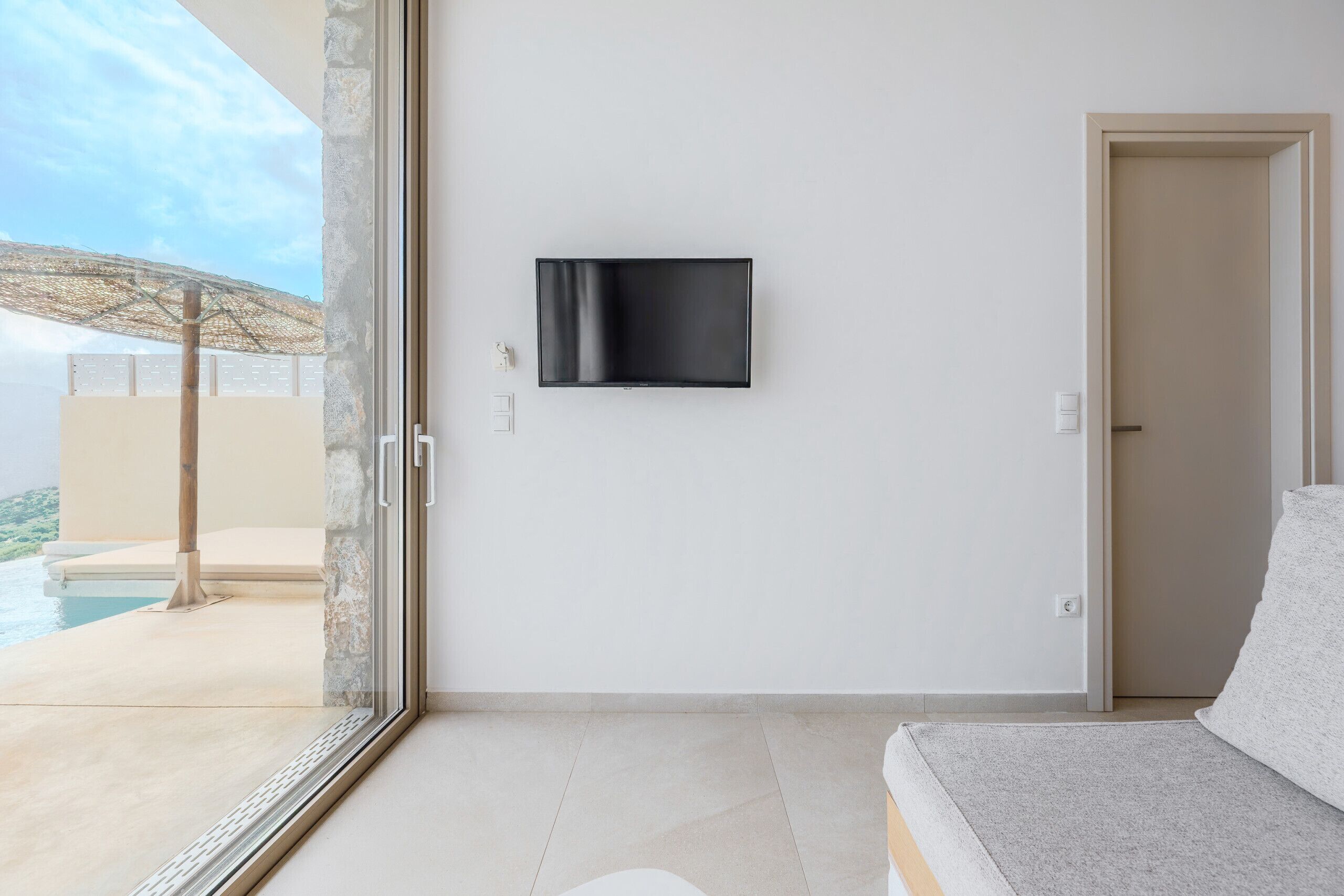 Une chambre d’hôtel moderne dotée d’une grande fenêtre, d’un téléviseur à écran plat fixé au mur, d’une porte et offrant une vue sur une piscine et des parasols.