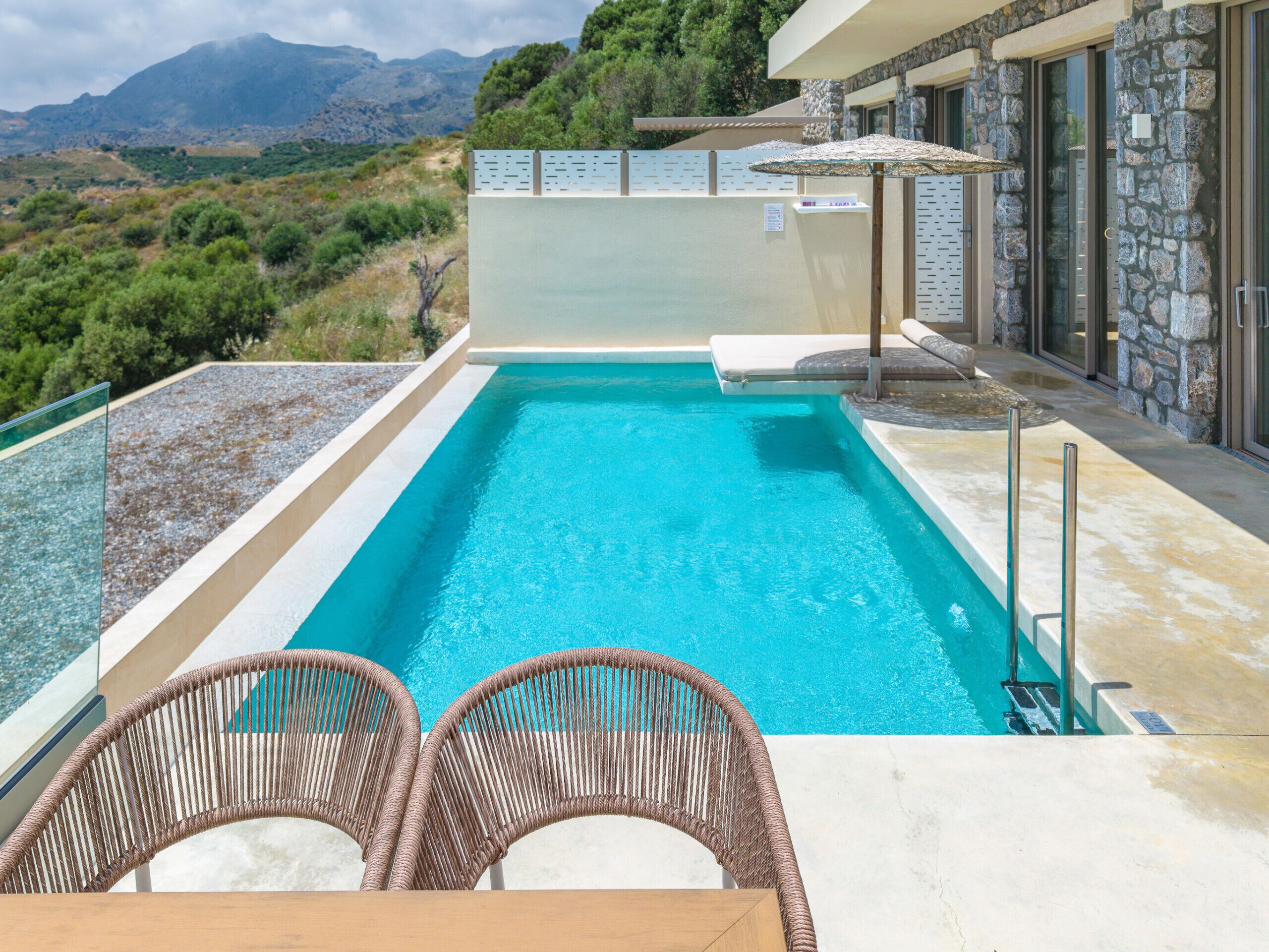 Ein rechteckiger Swimmingpool mit klarem, blauem Wasser, umgeben von Steinmauern und einer Glasbrüstung. Im Vordergrund stehen Korbstühle und ein Tisch, im Hintergrund ein Steinhaus mit Glastür und Fenstern.