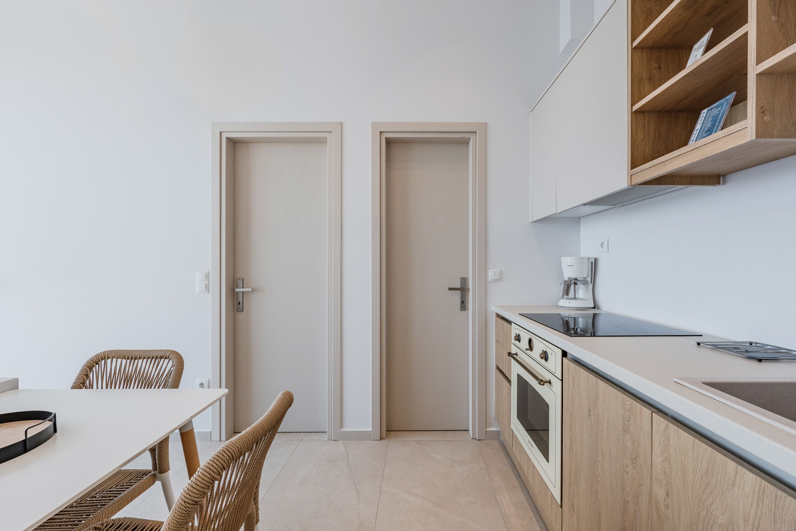 A modern kitchen with a dining area, featuring a white table, wicker chairs, wooden cabinets, and a coffee maker.