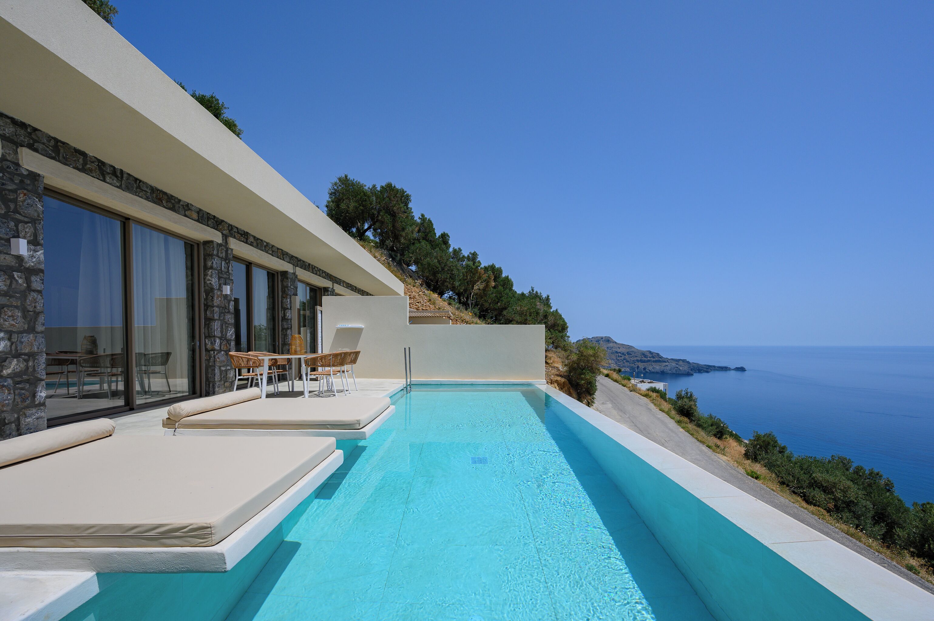 Un área moderna junto a la piscina con vista despejada al mar, paredes de piedra y un espacio de descanso con sillas y una mesa.