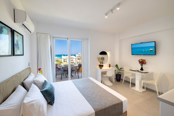 A hotel room with a bed, a desk, a chair, a TV, and a view of the sea.