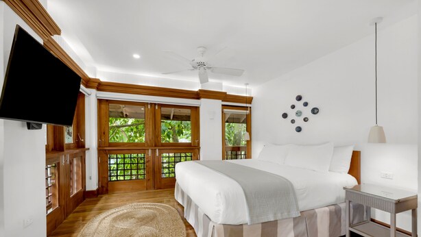 A bedroom with a large bed, wooden flooring, a ceiling fan, a TV, and a view of greenery through the windows. - Idle Awhile Beach - Family Suite in Negril (Negril)