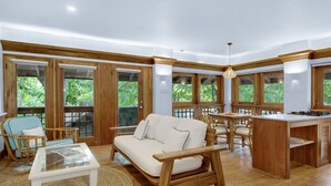 A spacious living area with a sofa, dining table, and chairs, large windows with a view of greenery, and a kitchen area with wooden cabinetry. - Idle Awhile Beach - Family Suite in Negril (Negril)
