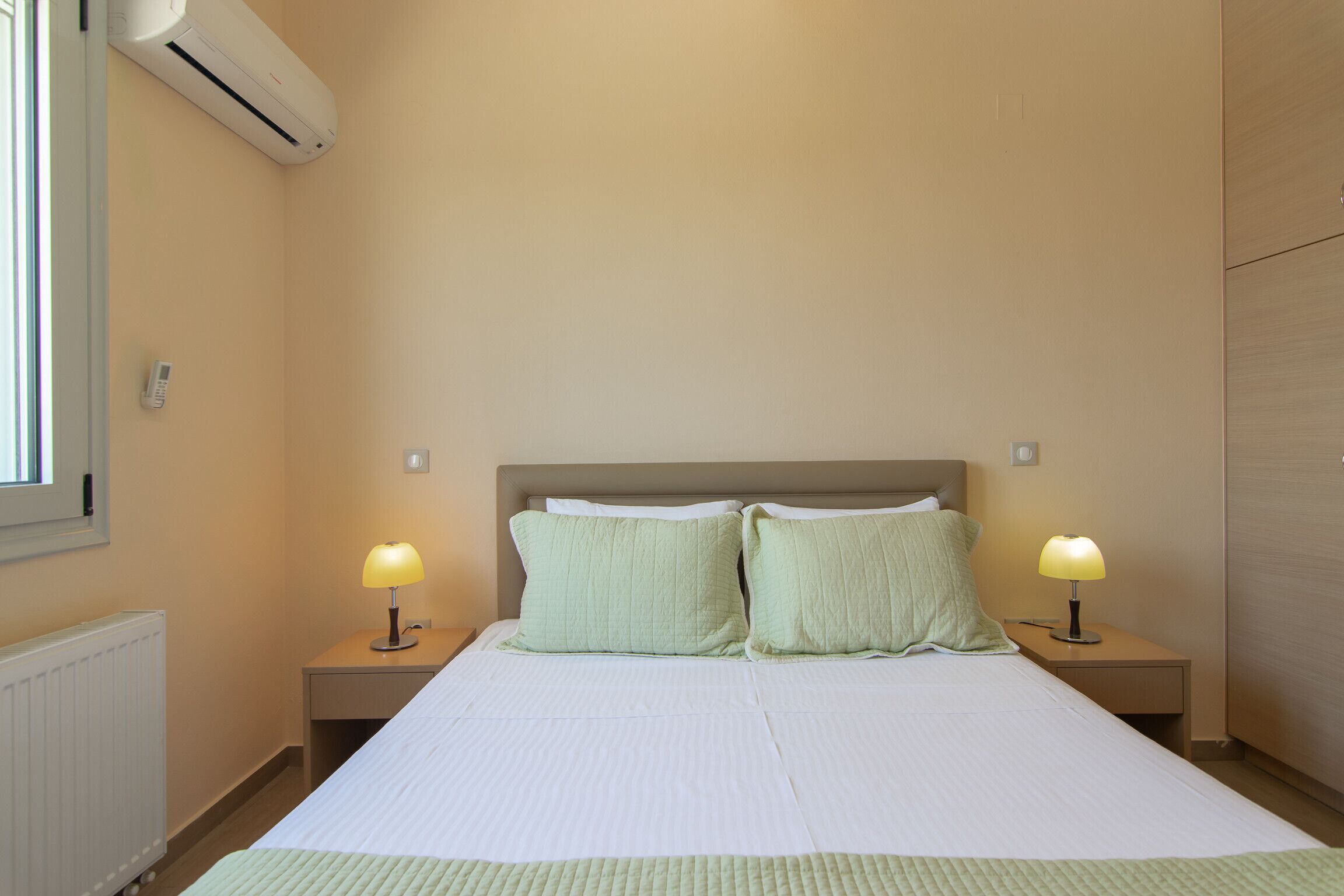 A neatly made bed with two pillows, two bedside lamps, and an air conditioning unit on the wall.
