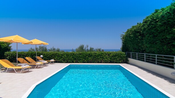 A rectangular swimming pool with clear blue water, surrounded by yellow lounge chairs and umbrellas, set against a backdrop of greenery and a clear sky. - Yanni's villas (Rethymno)