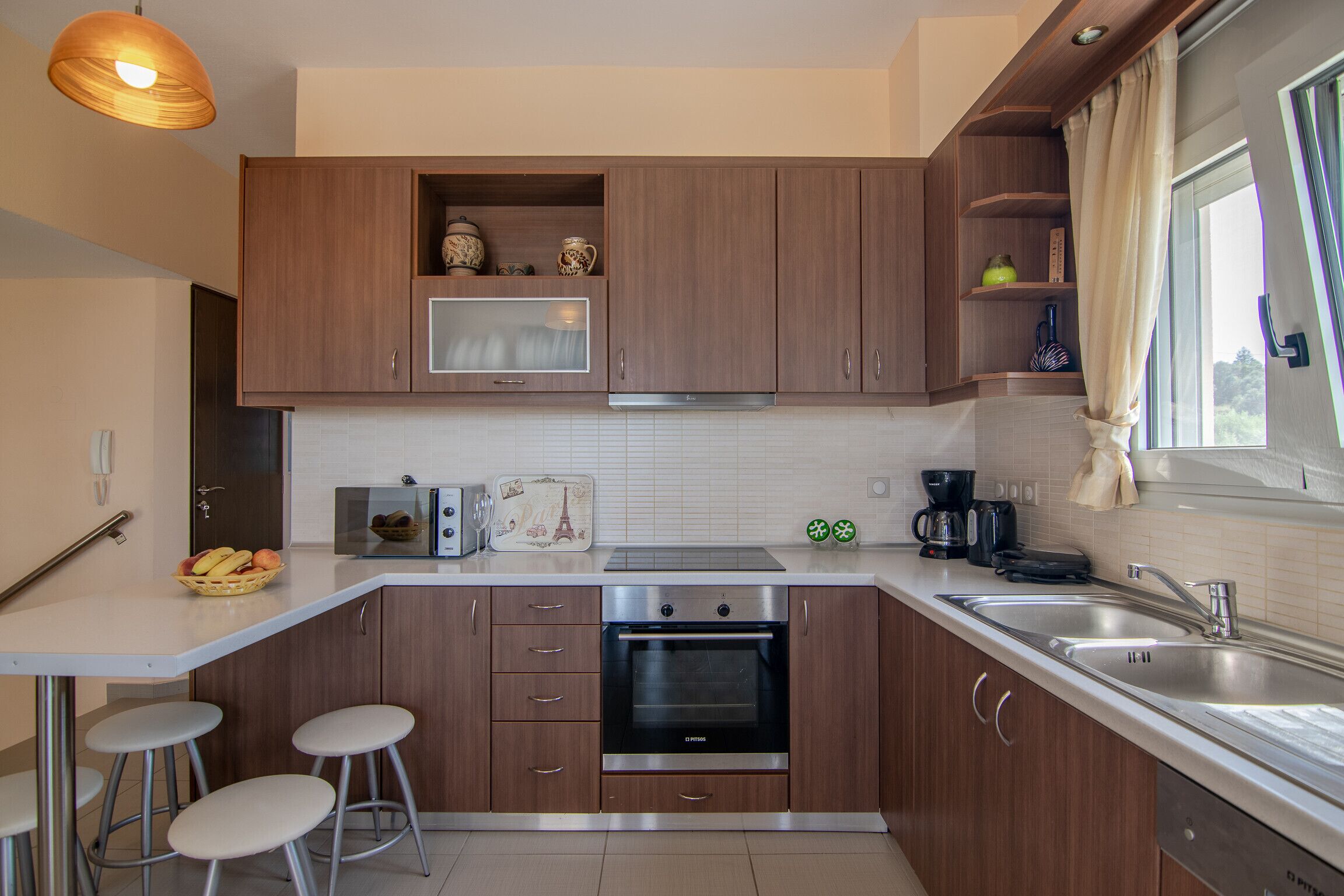 Una cocina moderna con armarios de madera, horno empotrado, microondas y pileta de acero inoxidable. Hay taburetes en la barra y una ventana con cortinas.