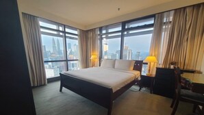 A hotel room with a large bed, two bedside lamps, a desk, and a chair. The room has large windows with curtains and a view of a city skyline. - 4-bedroom apartment in marvelous City with AC, fitness room (City)