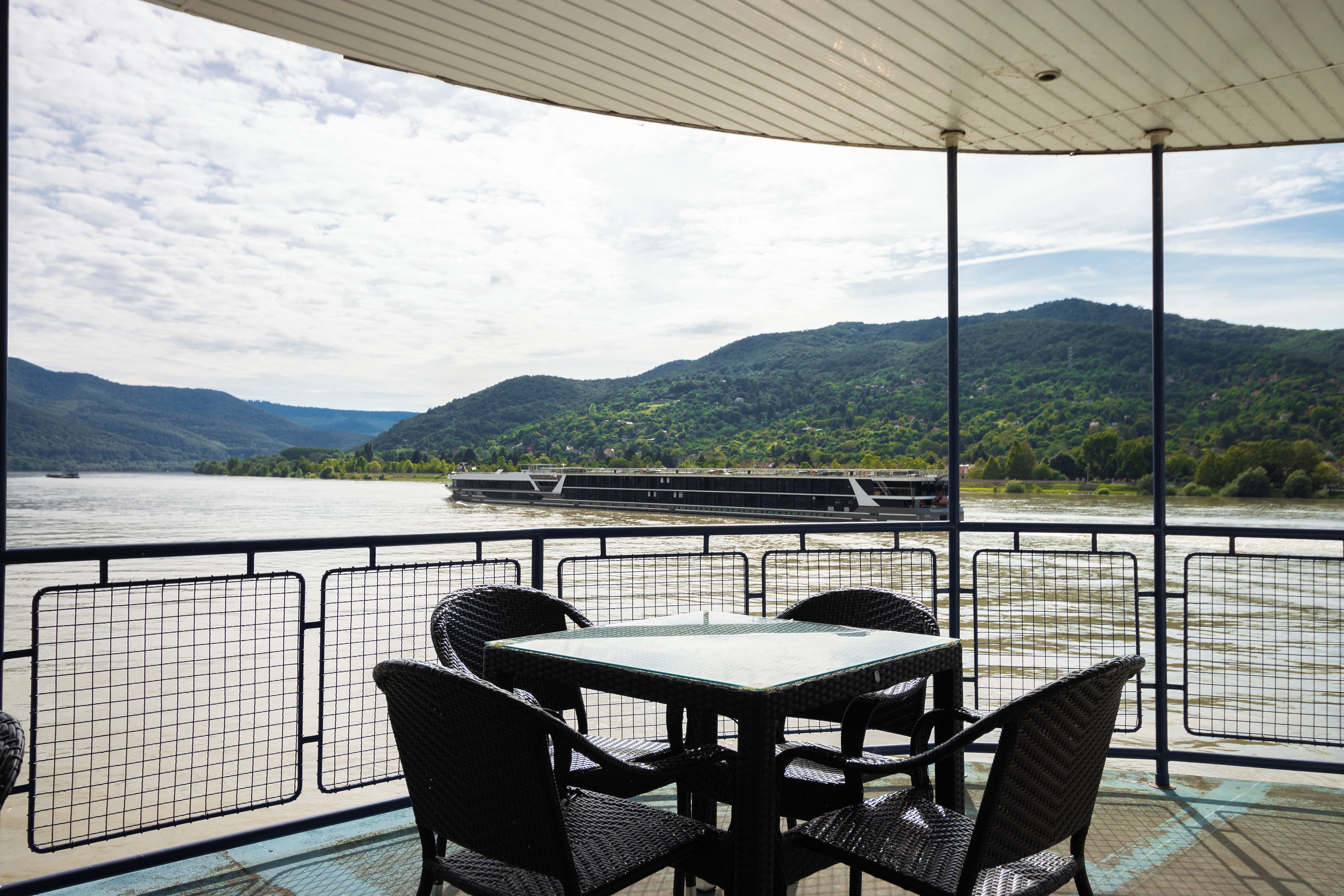 A table and chairs on a deck with a view of a river and mountains.