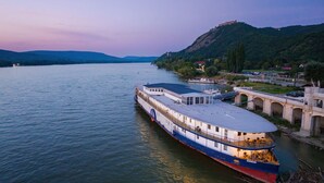 Un bateau fluvial doté d’un pont couvert et d’un espace repas, amarré près d’un pont et d’une colline sur laquelle se dresse une structure ressemblant à un château.