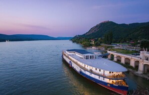 A riverboat with a covered deck and a dining area, docked near a bridge and a hill with a castle-like structure in the background. - Aquamarina Hotel Visegrád (Visegrád)