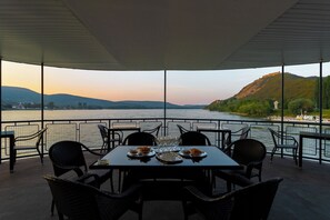 An outdoor dining area with a table set for four, overlooking a body of water and hills. - Aquamarina Hotel Visegrád (Visegrád)