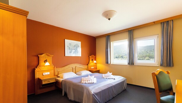 Une chambre d’hôtel avec un lit, deux tables de chevet avec des lampes, une chaise et une fenêtre avec des rideaux.