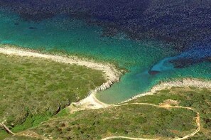 Oversigtsbillede af et kystområde med krystalklart turkis vand, en sandstrand og en smal vegetationstribe, der adskiller stranden fra vandet.