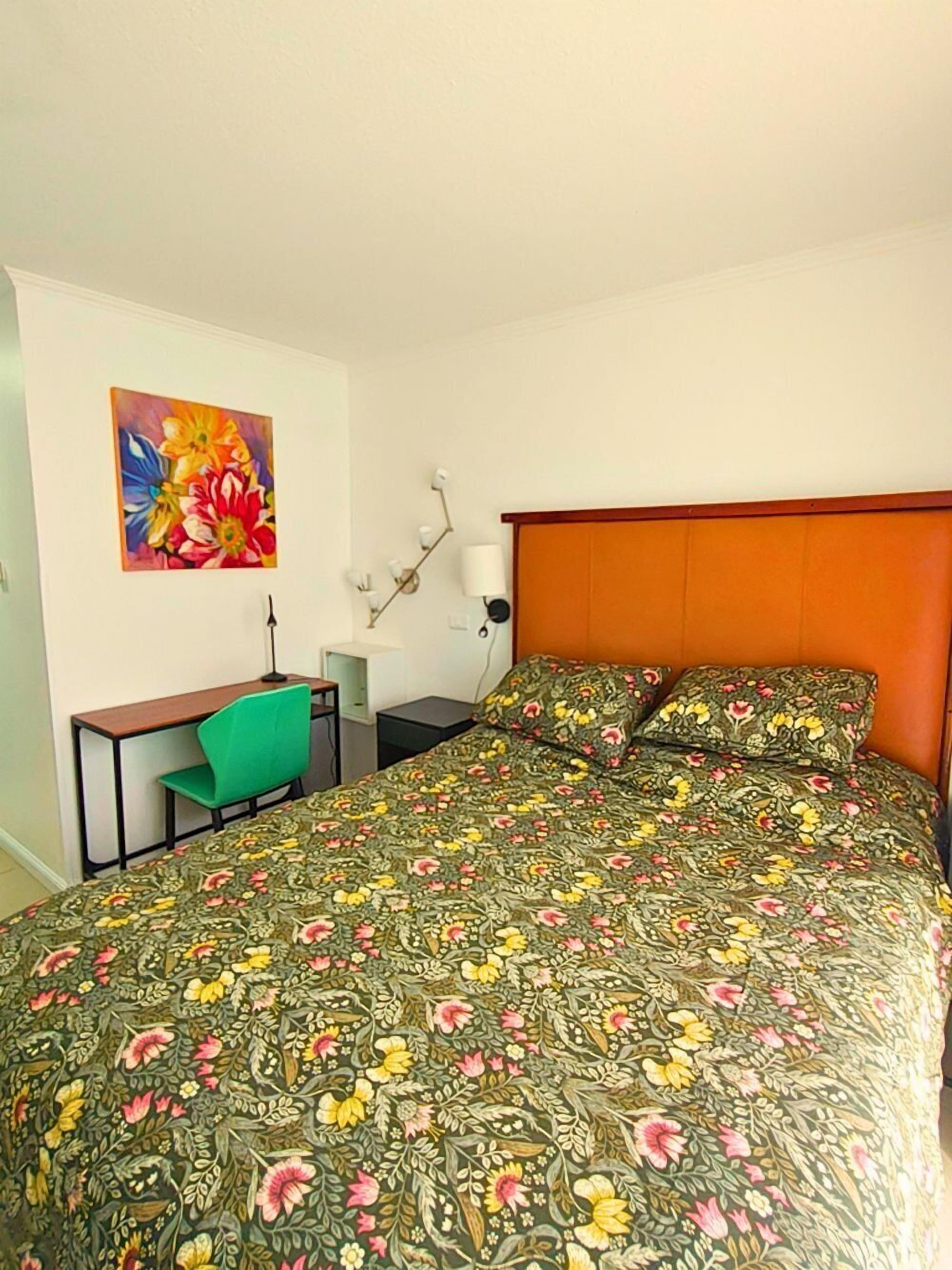 A bedroom with a floral bedspread, a green chair, a desk, and a colorful painting on the wall.