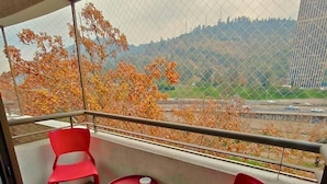 Un balcón con dos sillas rojas y una pequeña mesa, con vista a un paisaje urbano con follaje otoñal y un edificio distante.
