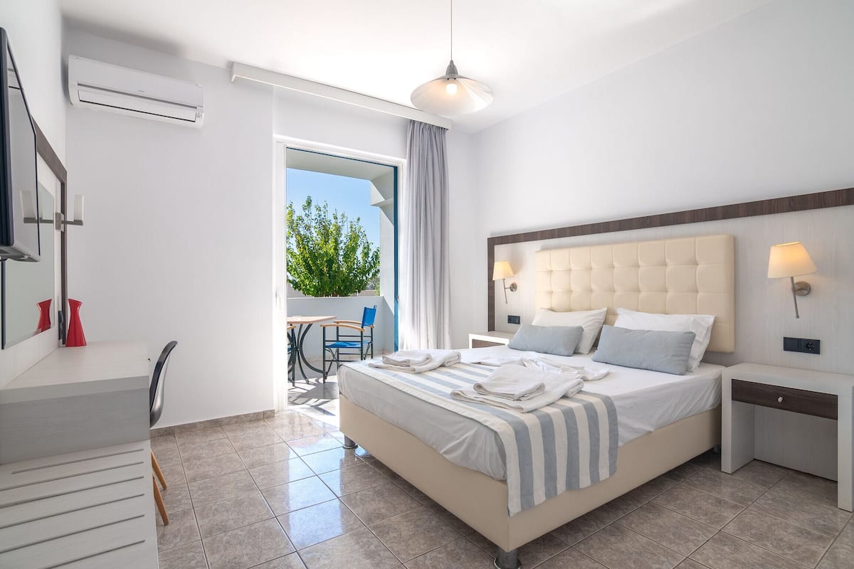 A modern hotel room with a large bed, a desk, and a balcony with a view of greenery.