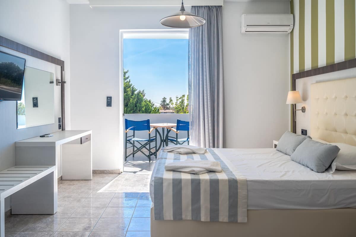 A modern hotel room with a bed, bedside table, and a desk with a chair. There is a balcony with a view of greenery and a clear sky.