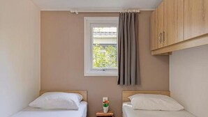 A small, single-room accommodation with two single beds, a window with curtains, and a wooden cabinet.