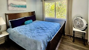 Een slaapkamer met een bed, nachtkastje, wandventilator en raam met gordijnen.
